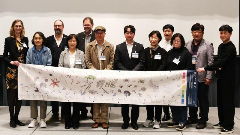 Speakers at the international symposium. © Young-Nam Kim/ KOEM. A group of people on a stage holding an educational banner with Wadden Sea plants and animals. © Young-Nam Kim/ KOEM.
